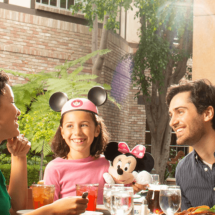 Mom, dad, and daughter wearing Mickey Mouse ears and clutching stuffed Minnie Mouse enjoy meal together on a vacation to Disneyland in Anaheim, California.